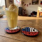 Dos posavasos Viva Chile impresos en 3D sobre una mesa de madera, uno bajo un vaso de terremoto, con banderas chilenas al fondo