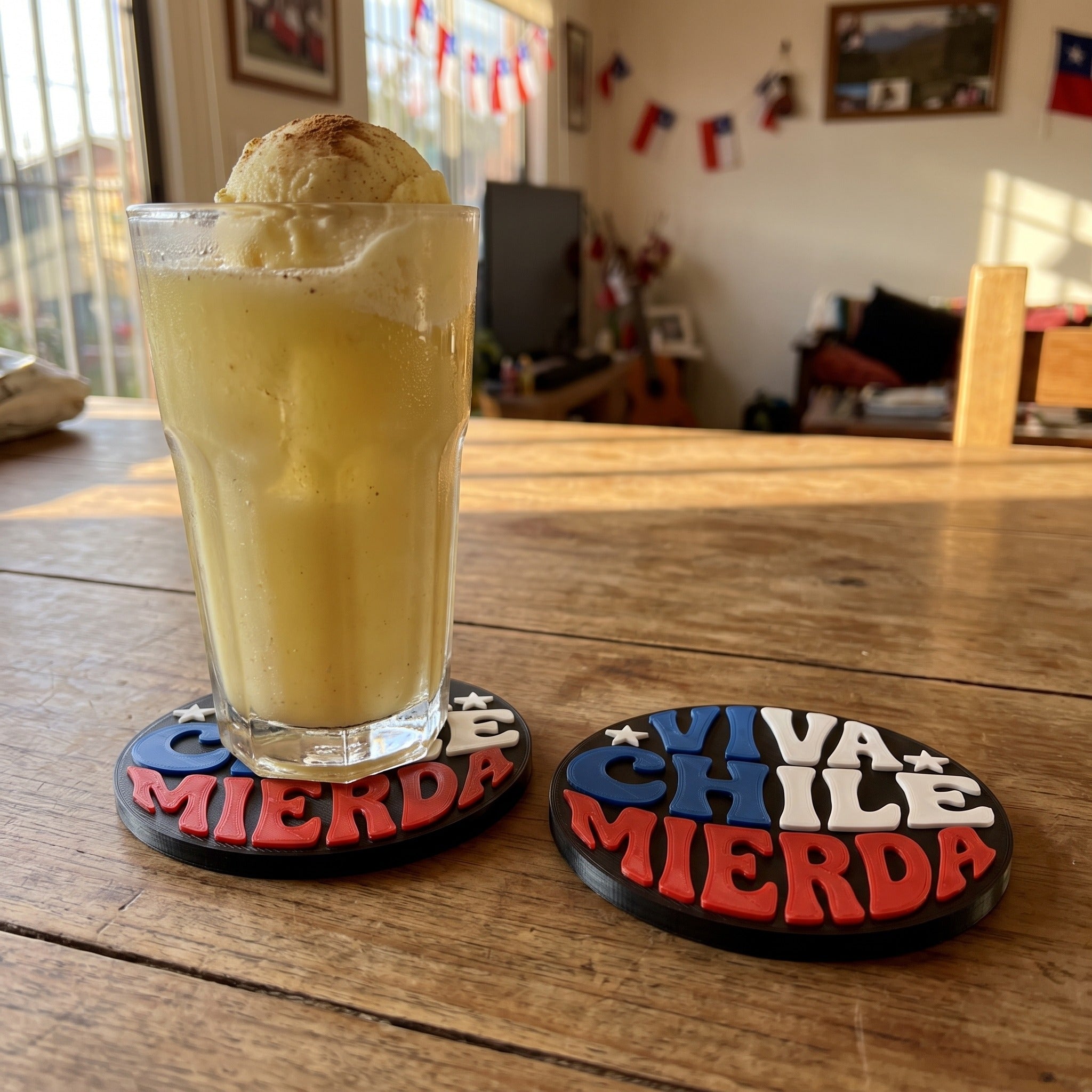 Dos posavasos Viva Chile impresos en 3D sobre una mesa de madera, uno bajo un vaso de terremoto, con banderas chilenas al fondo