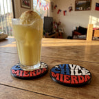 Dos posavasos Viva Chile impresos en 3D sobre una mesa de madera, uno bajo un vaso de terremoto, con banderas chilenas al fondo